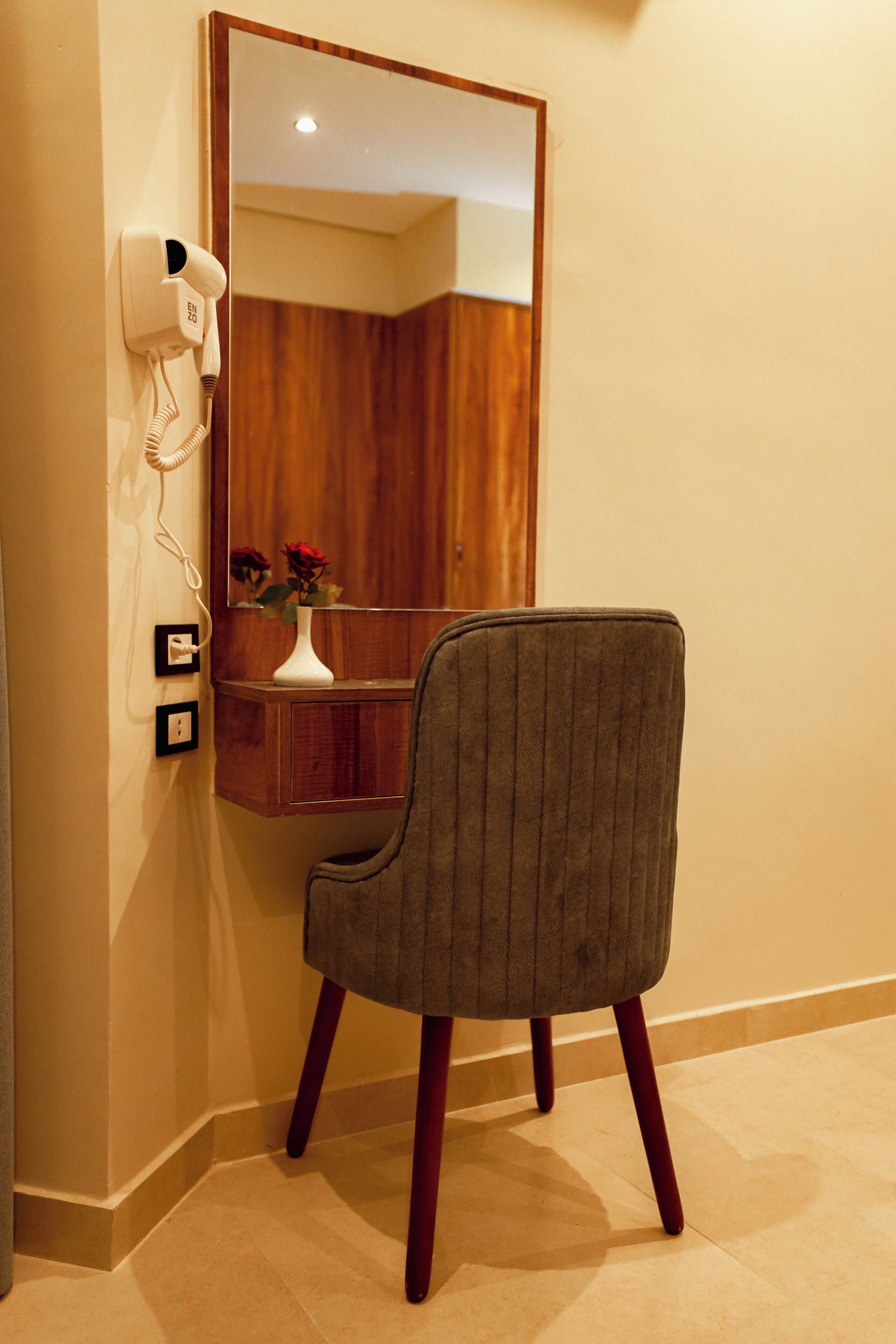 A room with a hairdryer mounted on the wall, a mirror, a wooden vanity, and a grey upholstered chair.