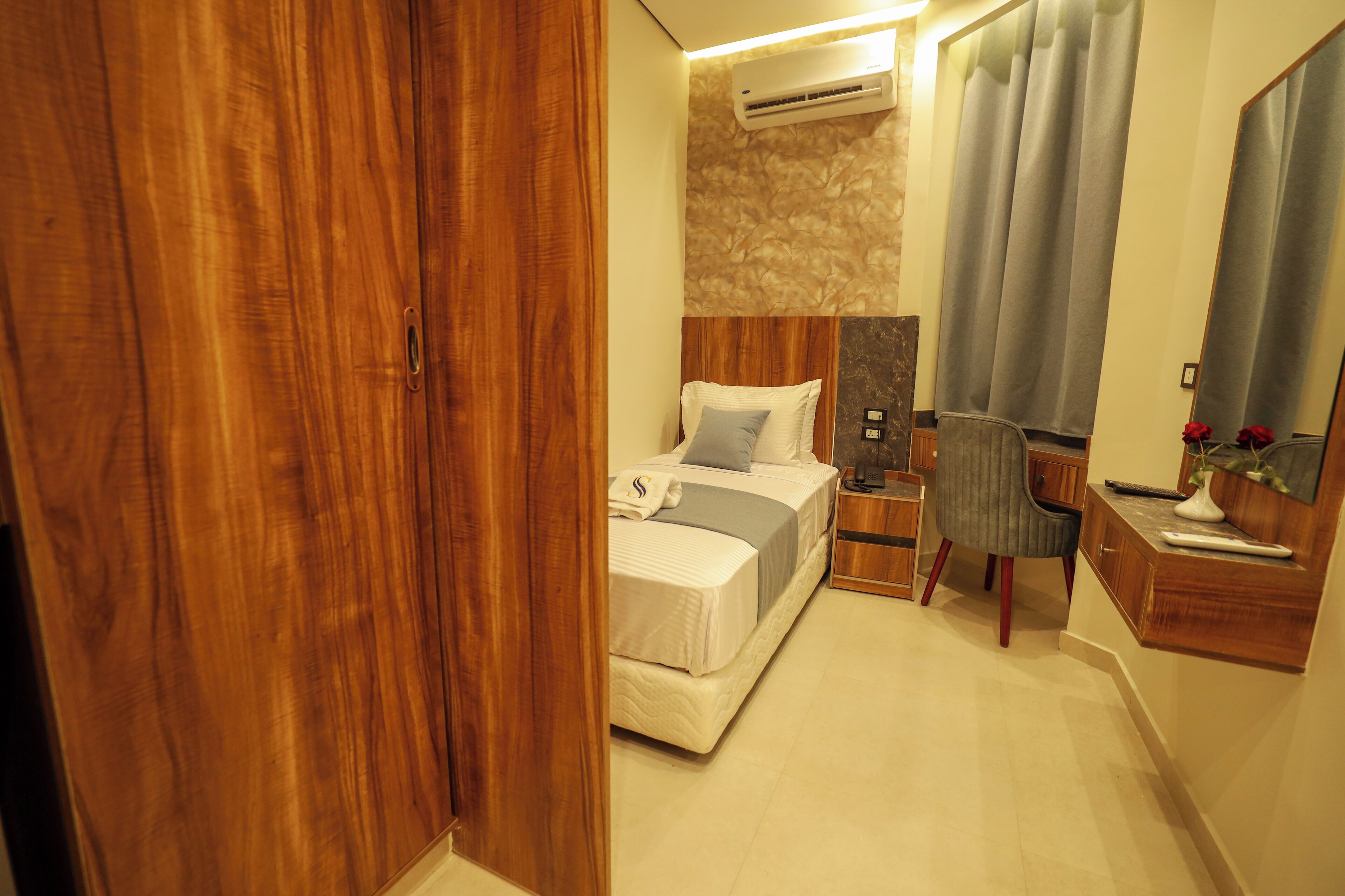 A bedroom with a wooden wardrobe, a bed with pillows, a nightstand, a chair, a desk with a mirror, and an air conditioning unit.