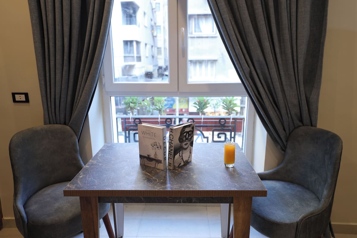 Una habitación con una mesa, dos sillas, un ventanal con cortinas y vistas a edificios y plantas al exterior.