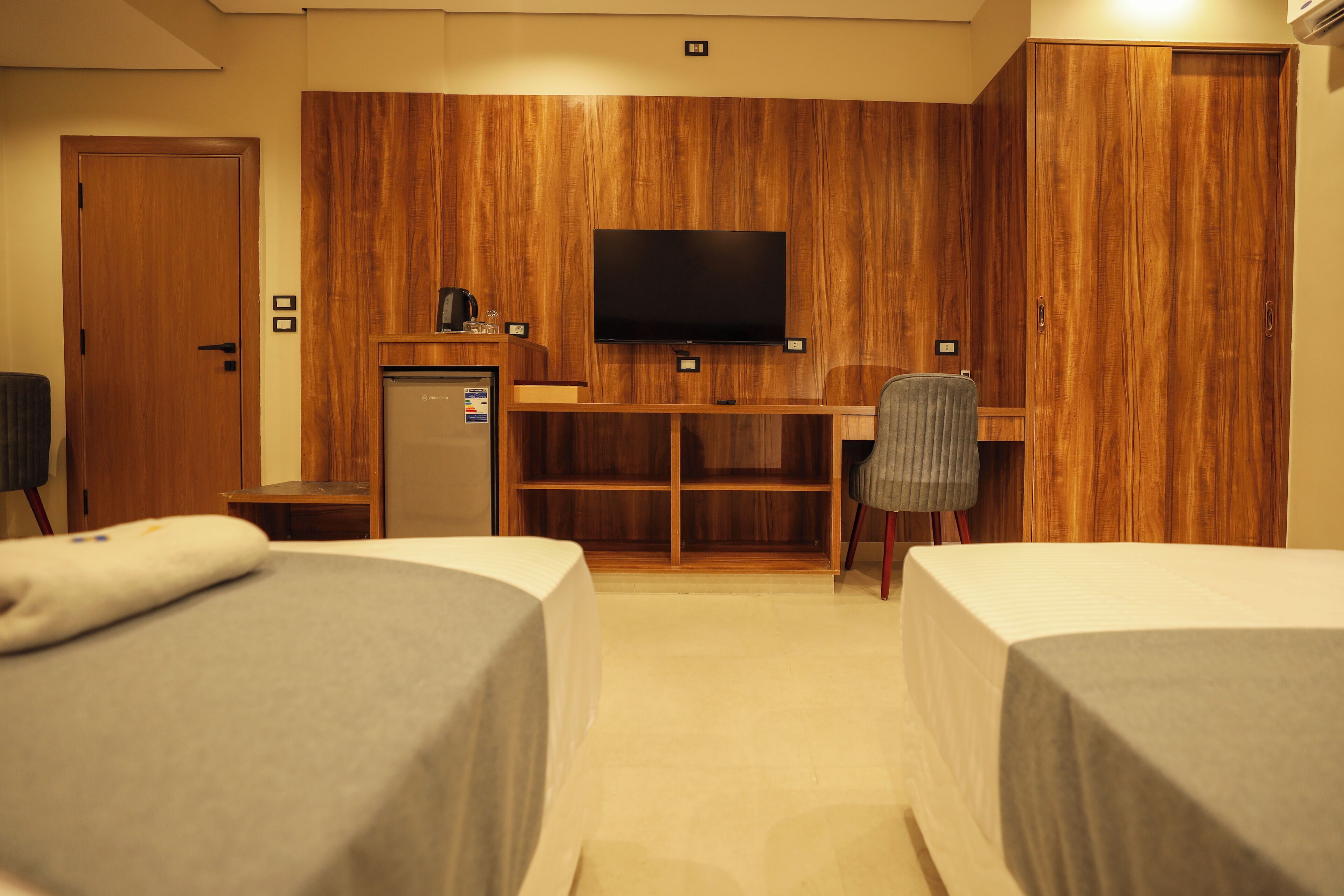 A hotel room with two beds, a desk with a TV, a refrigerator, and a chair.