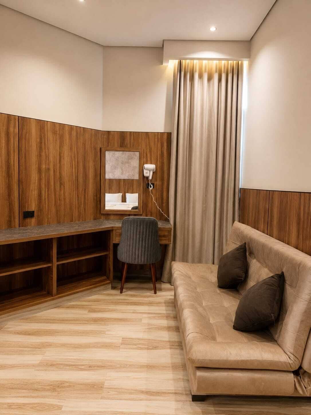 A modern room with a wooden desk, a grey chair, a beige sofa with cushions, and a large window with curtains.