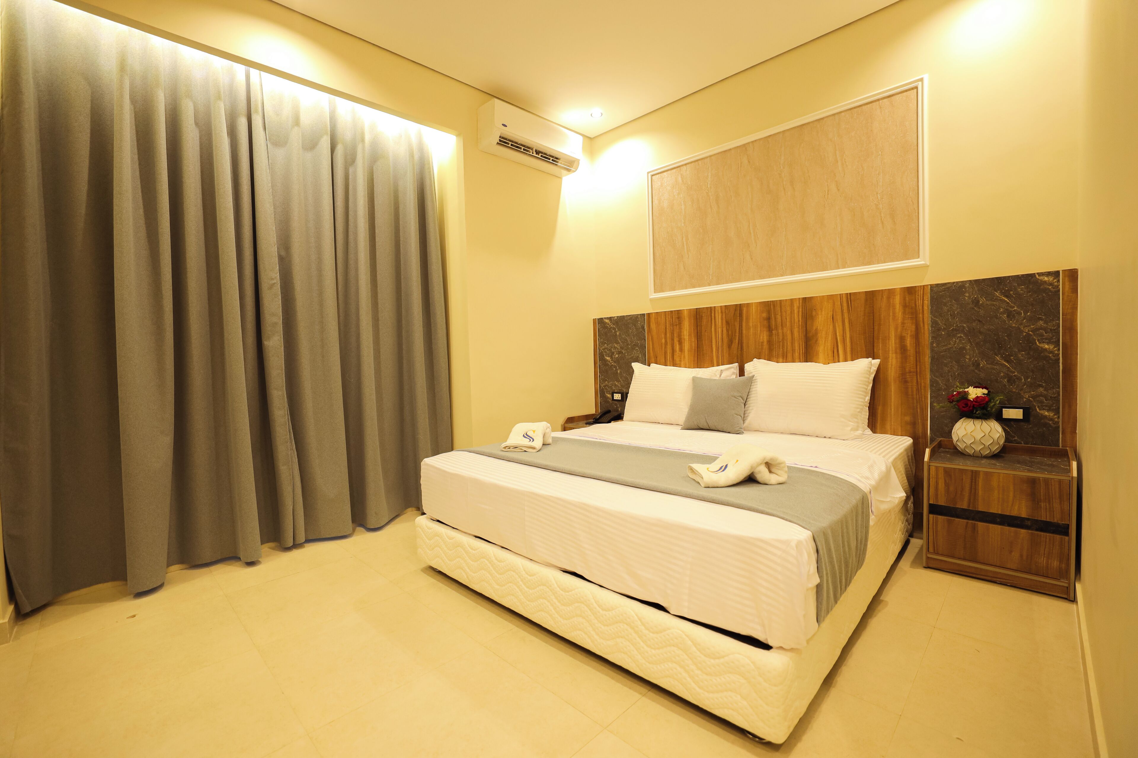 A hotel room with a large bed, a bedside table with a vase, and a wall-mounted air conditioner.