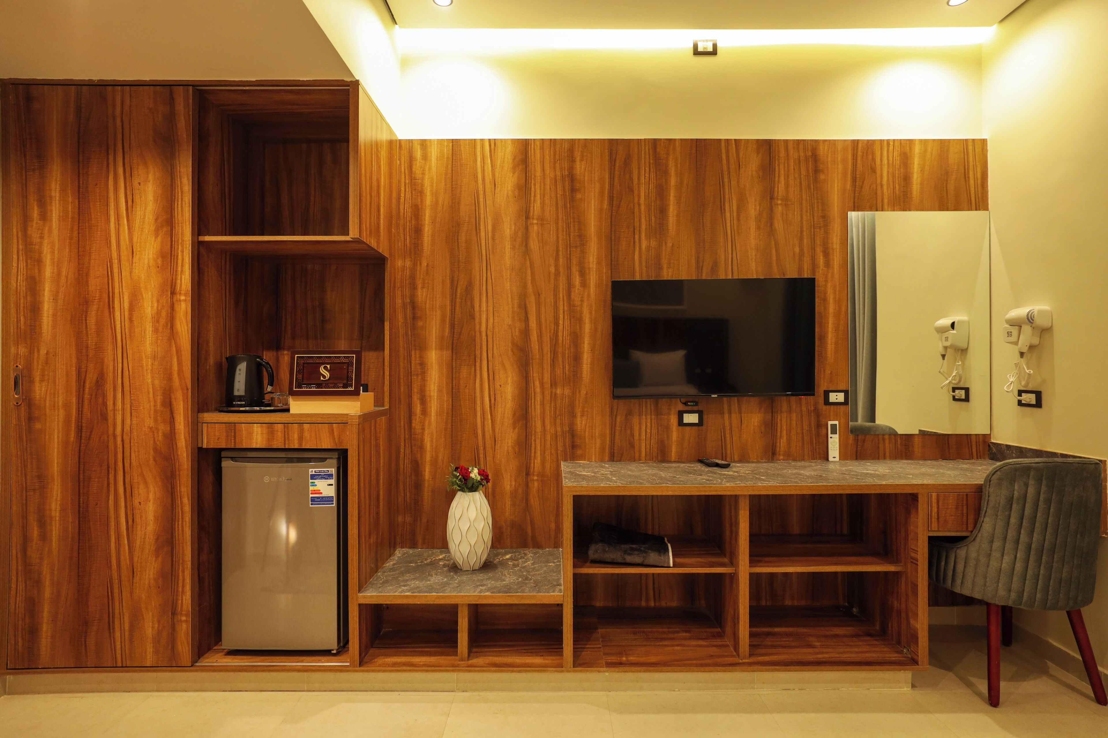 Une chambre d’hôtel compacte avec des panneaux de bois, un téléviseur intégré, un petit réfrigérateur, une coiffeuse avec un miroir et un coin salon avec une chaise.