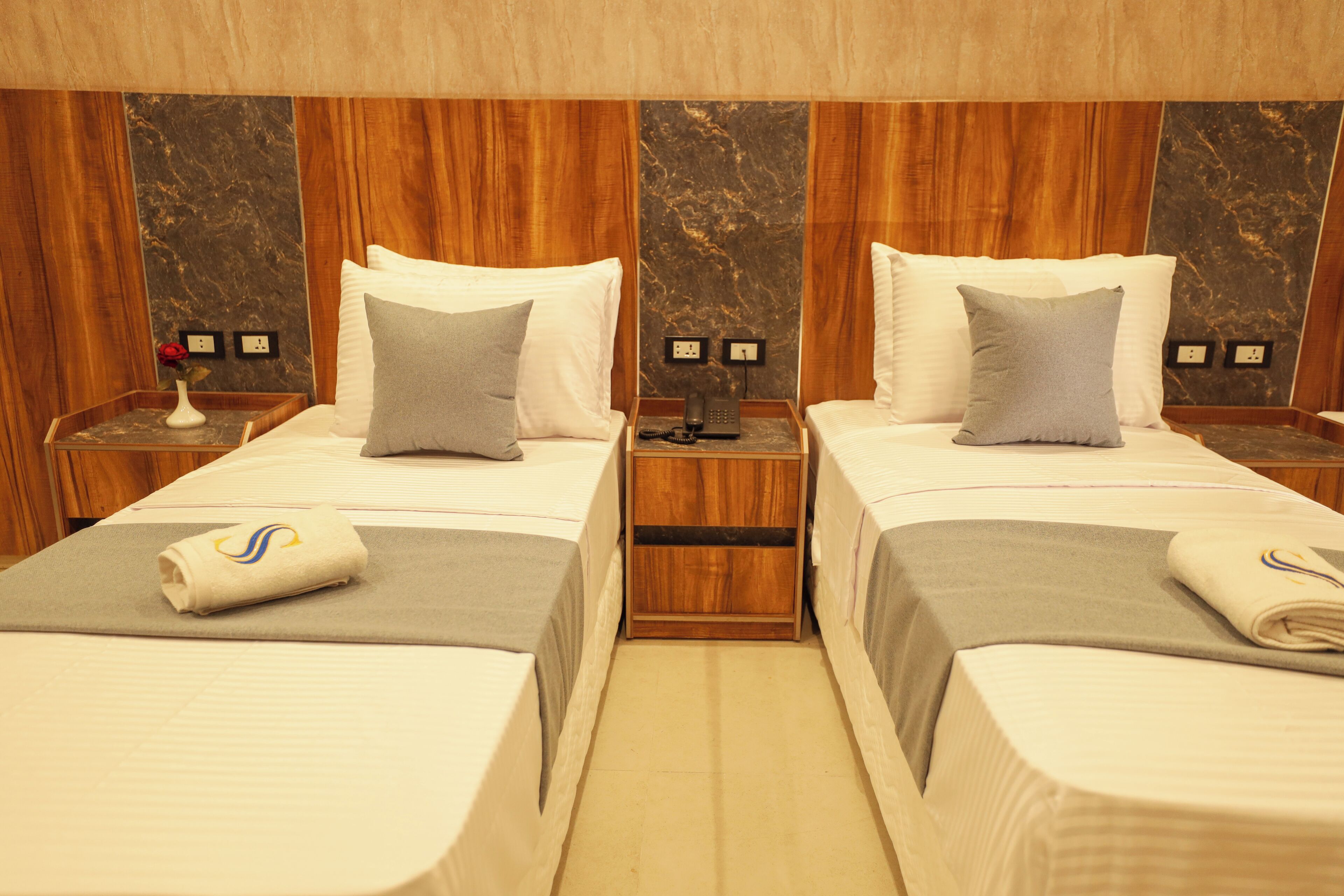 A hotel room with two beds, wooden headboards, marble wall panels, bedside tables with lamps, and a small vase with a flower on one table.