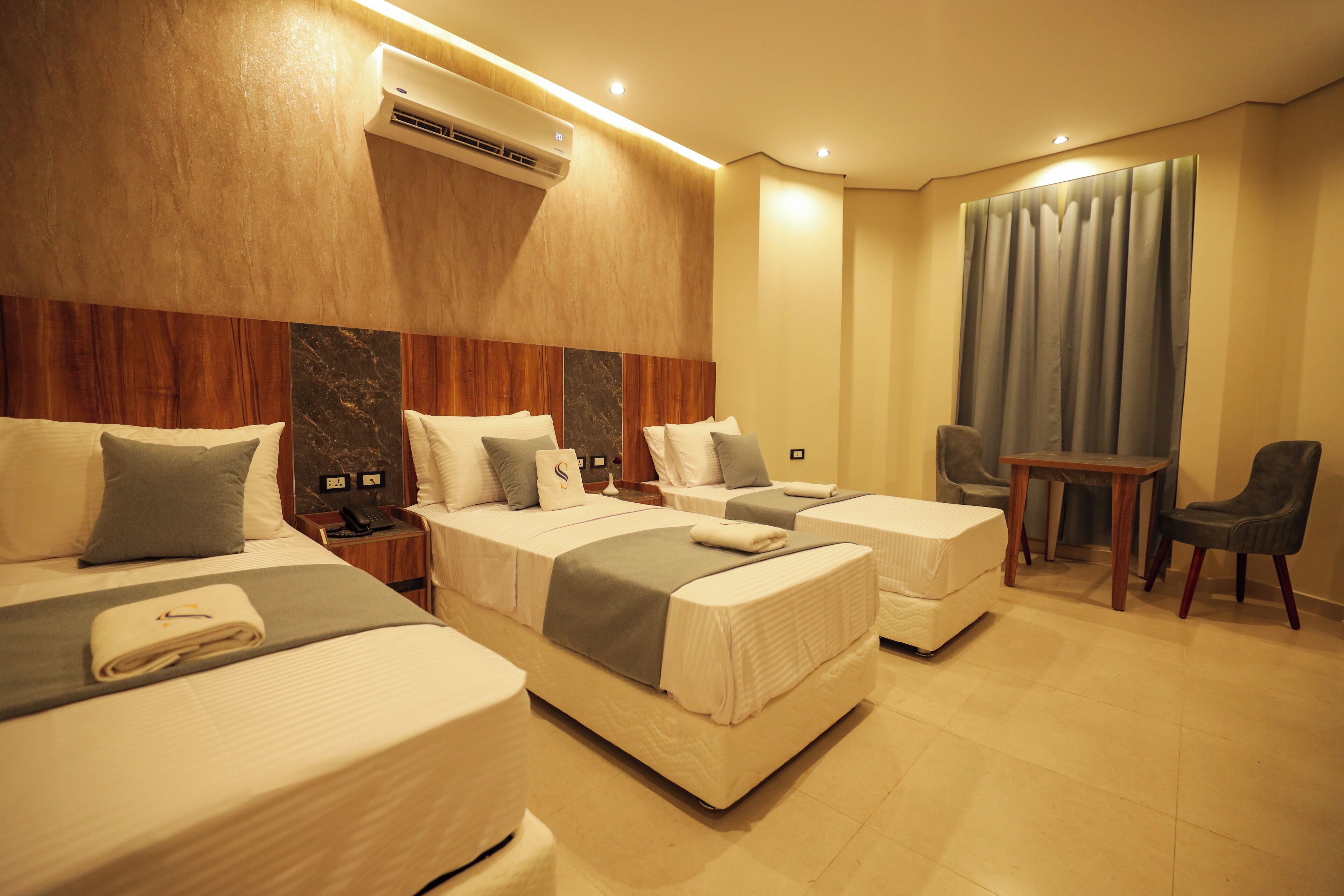 Quarto de hotel com três camas, uma escrivaninha, uma cadeira e uma janela grande com cortinas.