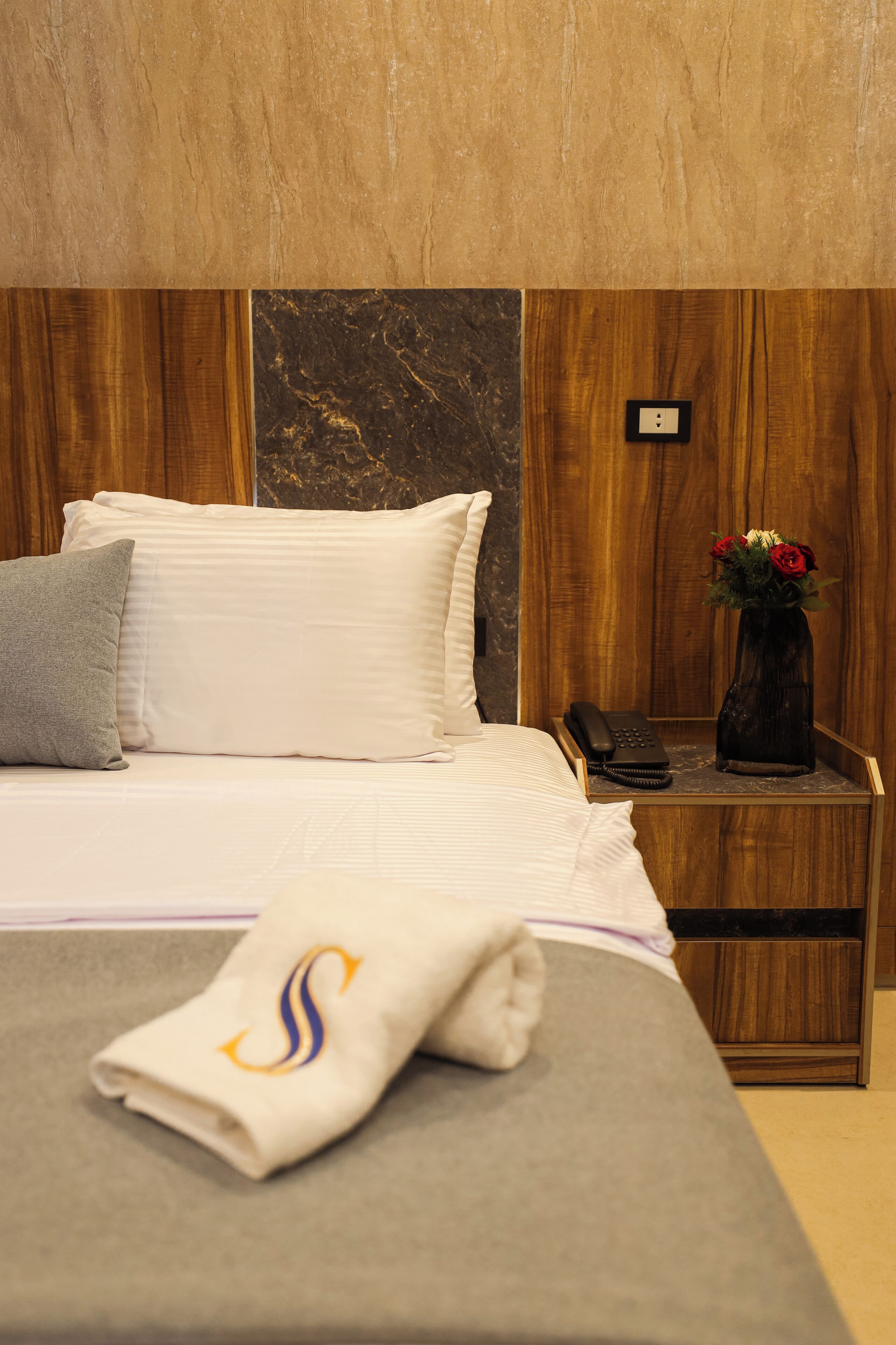 A neatly made bed with white linens and a gray pillow, a wooden nightstand with a phone and a vase of flowers, and a wall with a marble-like texture.