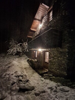 Ein Steinhaus mit geneigtem Dach und einer Holztür. Schnee liegt auf dem Boden und um das Gebäude herum.