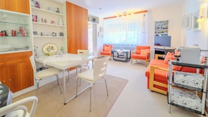 A modern living room with a dining area, featuring a white table, orange chairs, and a TV. - FE-WO Paradiso, 78141 Schönwald in the Black Forest With swimming pool and sauna 1000m.a.s.l. (Schönwald im Schwarzwald)