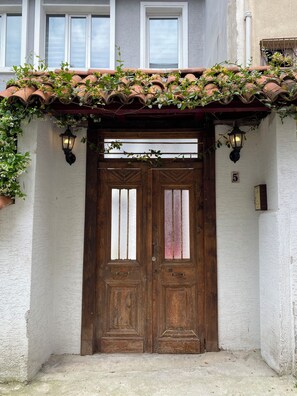 Une porte à deux battants en bois, adossée à un mur blanc et surmontée d’un toit recouvert de tuiles.