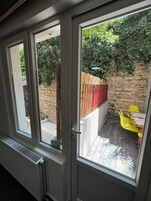 Una habitación con una puerta de vidrio corredera que da a un patio con una cerca roja, un muro de piedra y una silla amarilla.