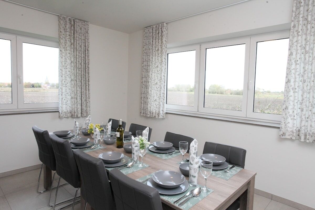 Une salle à manger avec une table dressée pour quatre personnes, de grandes fenêtres avec des rideaux et une vue sur l’extérieur.