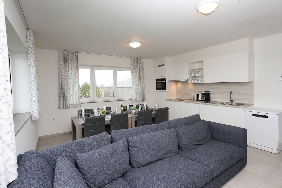 Un appartement moderne avec un canapé gris, un coin repas, une cuisine dotée de meubles blancs et une fenêtre avec des rideaux.