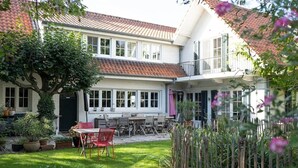 A two-story house with a balcony, a garden with a table and chairs, and a tree.