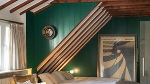 A cozy bedroom with a sloped ceiling, a bed, two wicker chairs, and a framed artwork on the wall.