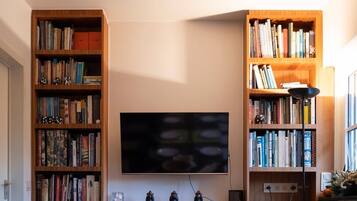 A dining area with a long table set for six, flanked by two wooden bookshelves filled with books, a flat-screen TV mounted on the wall, and a ceiling fan above.