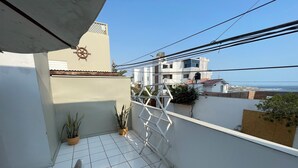 Varanda com piso de azulejos brancos, plantas em vasos, uma cadeira branca e vista para o mar e edifícios ao longe.