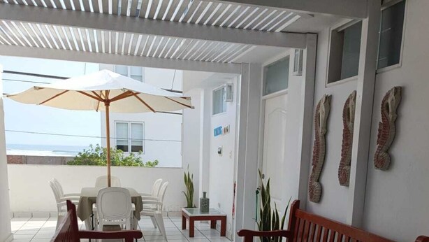Uma área de estar coberta ao ar livre, com mobiliário de madeira, uma mesa de vidro e vista para o mar.