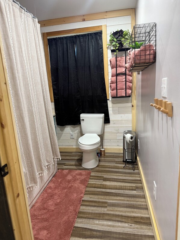 Une petite salle de bain avec des toilettes, un rideau de douche, un tapis rose et une étagère en métal où sont posées des serviettes.