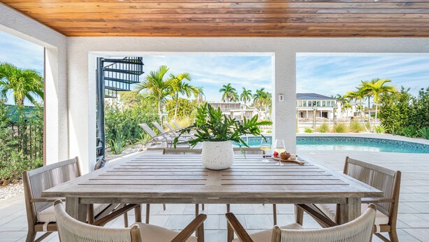 Un patio techado con una mesa y sillas de madera, una planta en maceta y vistas a una piscina y palmeras.