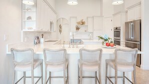 Una cocina moderna con armarios blancos, una isla grande con taburetes de bar, electrodomésticos de acero inoxidable e iluminación colgante.