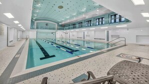 Ein großes Hallenbad mit mehreren Bahnen, einer hohen Decke mit runden Leuchten und einem Balkon in einer zweiten Ebene.