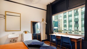 Habitación de hotel con cama, escritorio, silla, lavamanos y ventana con vistas a edificios.