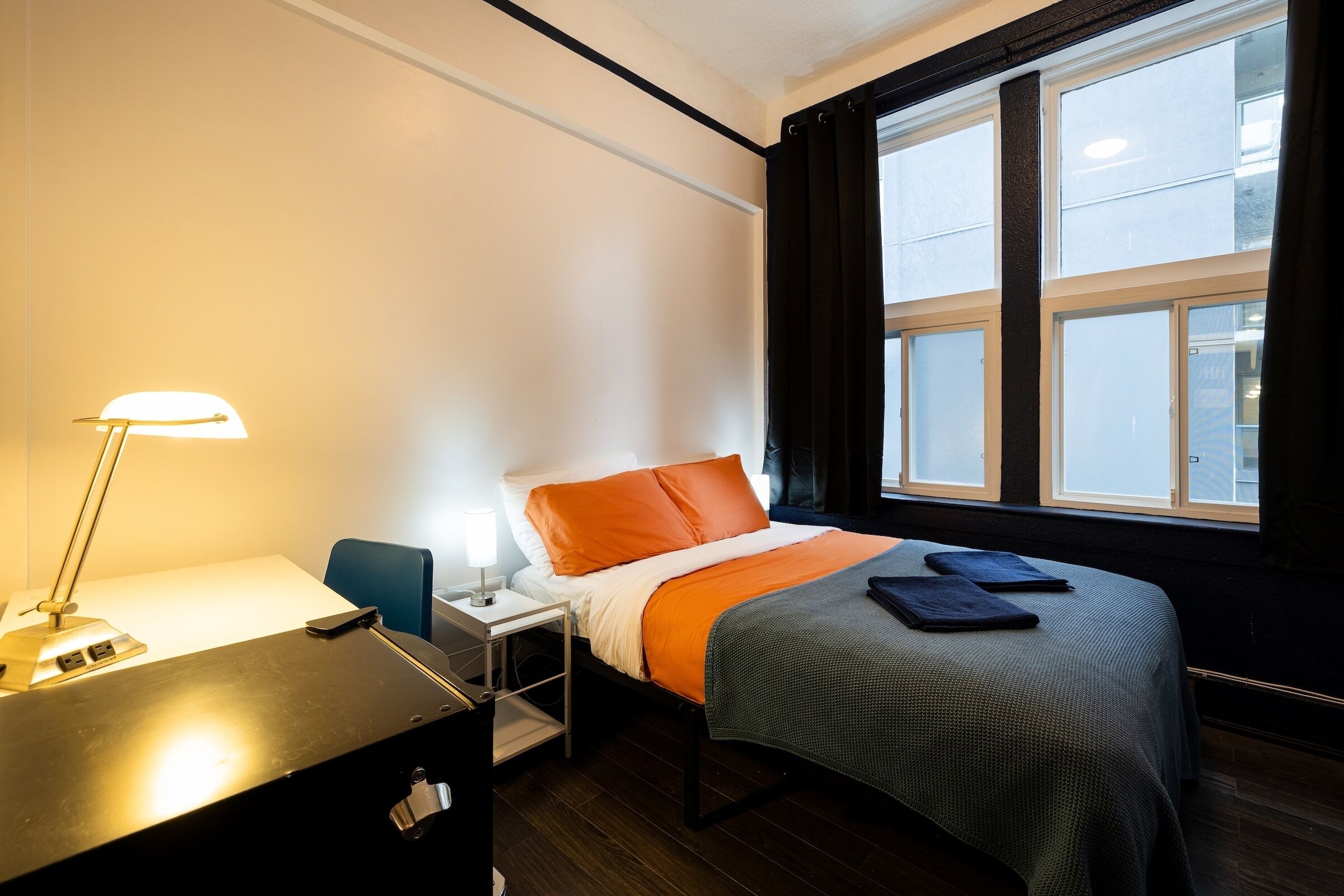 A hotel room with a bed, a desk with a lamp, a chair, and a window with curtains.