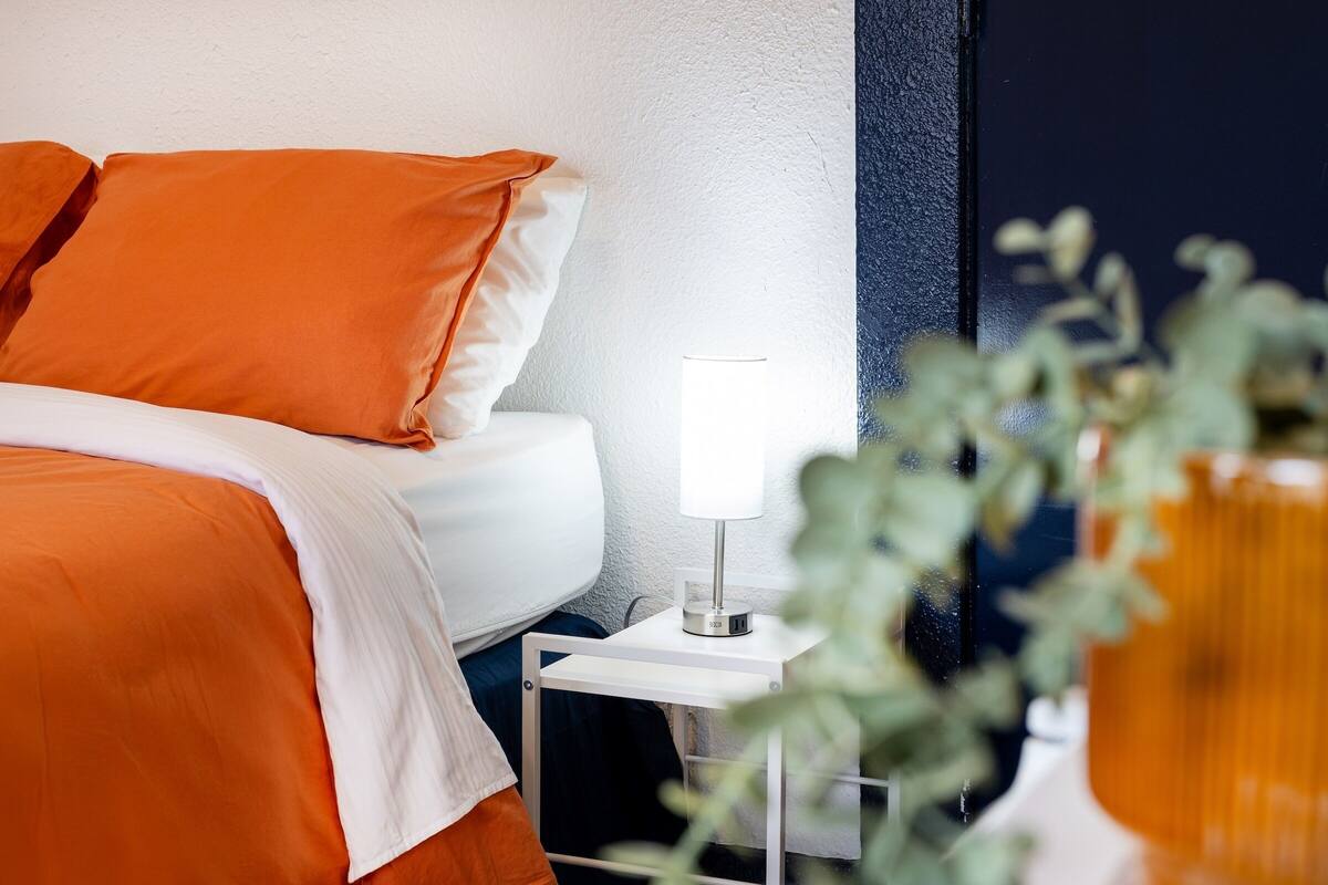 Een net opgemaakt bed met oranje beddengoed, een witte nachtkastje met een lamp en een plant op de voorgrond.