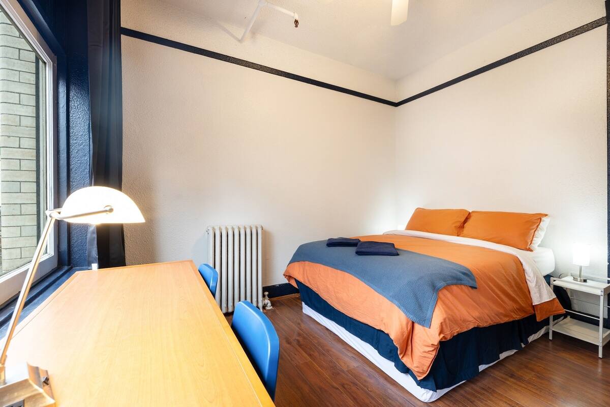 Een slaapkamer met een houten bureau, een bed met oranje en blauwe beddengoed, een nachtkastje en een raam met uitzicht.