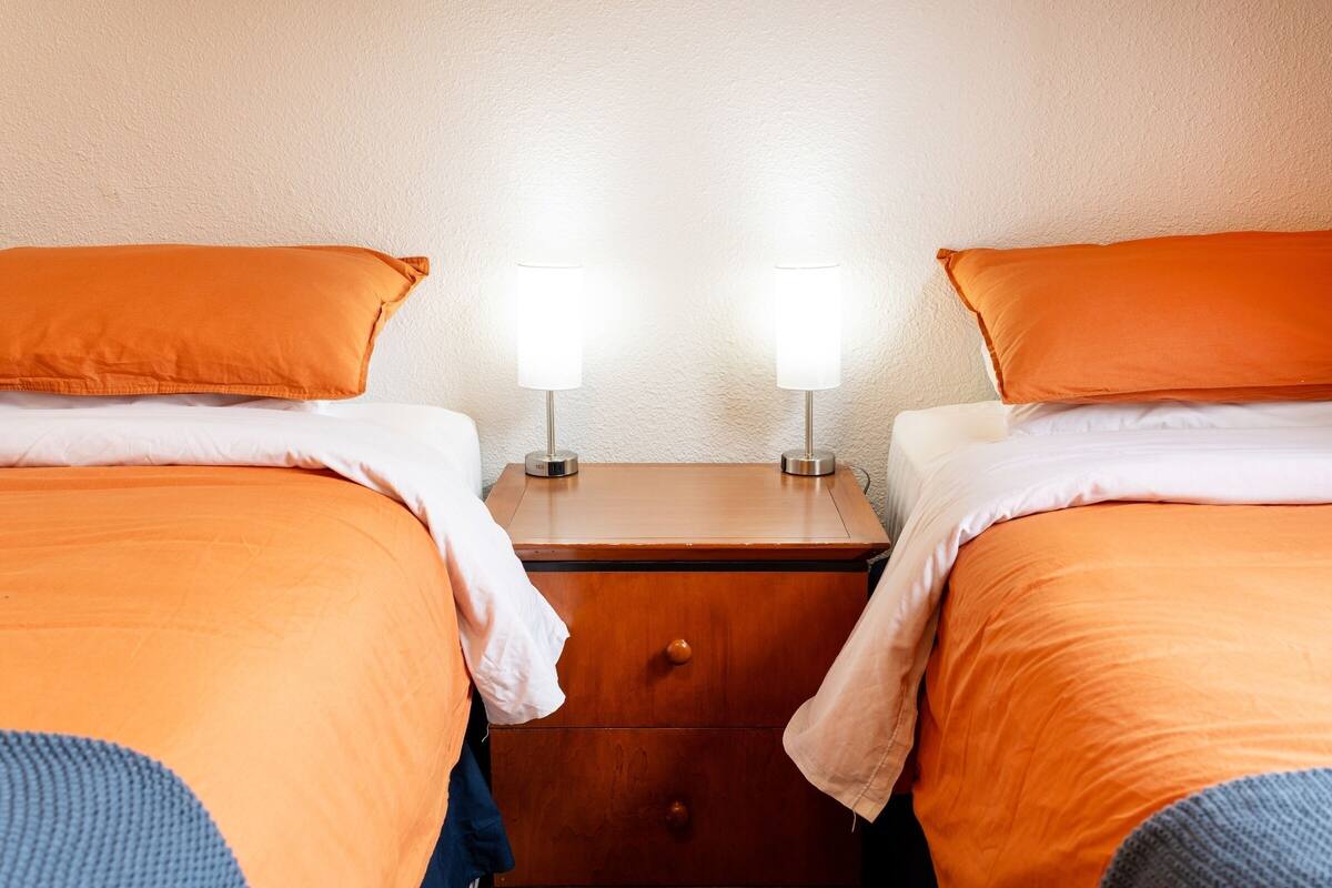 Twee bedden met oranje beddengoed, een houten nachtkastje met twee lampen en een witte muur.