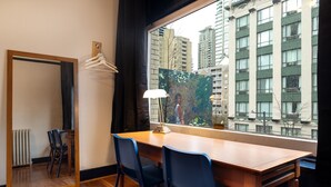 Habitación de hotel con escritorio de madera, dos sillas, un espejo, una ventana con vistas a edificios altos y un radiador.