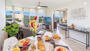 A breakfast setup with a variety of fruits and pastries on a table, a television, and a view of the outdoors through large windows. - Oceanview Retreat Walk to Beach & Restaurants (Kihei)