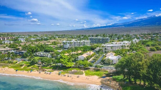 A coastal resort with beachfront properties, a central building complex, and a mountain in the background. - Beachfront Studio | Walk to Kamaole Beach + Pools (Kihei)