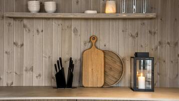 A kitchen counter with a wooden cutting board, a knife block, a lantern, and a shelf with bowls and a candle.