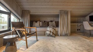 A cozy cabin interior with a wooden ceiling, a sofa with cushions, a chair, a TV, and a large window.
