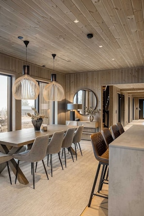 A modern interior with a wooden ceiling, a dining area with a long table and chairs, and pendant lighting.