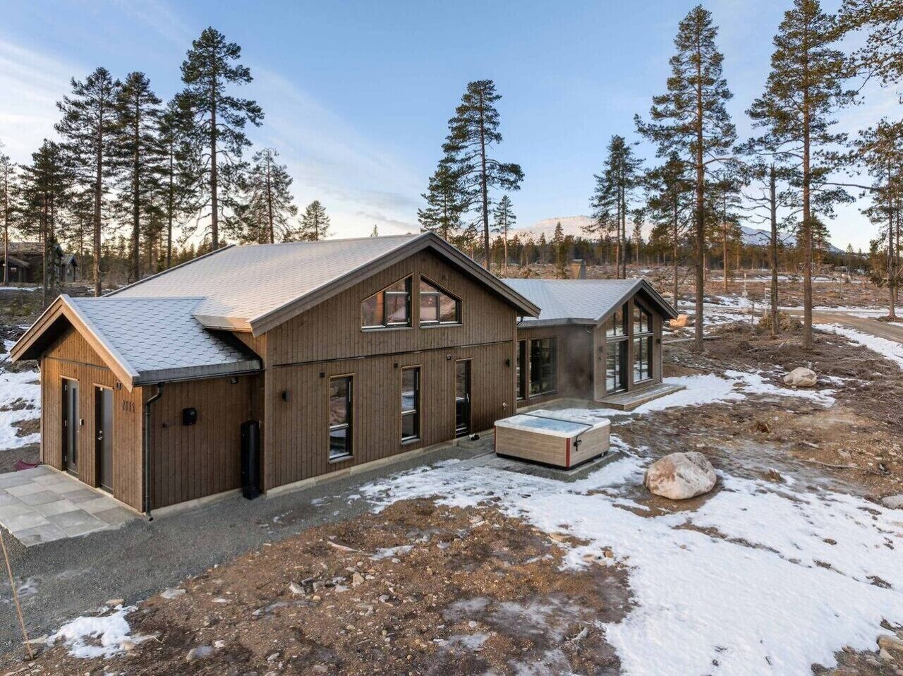 A two-story wooden cabin with a sloped roof, a large deck, and a surrounding forest.