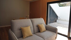 Een moderne woonkamer met een grijze bank, twee beige kussens, een bijzettafeltje met een vaas en een groot raam met uitzicht op een tuin.