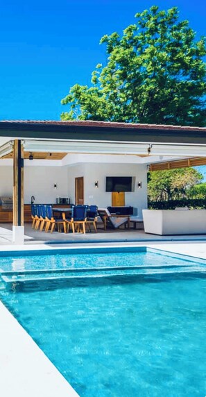 Un área de piscina moderna al aire libre con un espacio de estar cubierto y una piscina de color azul cristalino.