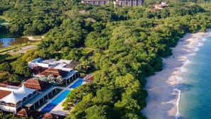 Vista aérea de un complejo turístico costero con playa, bosque denso y un gran conjunto de edificios al fondo.