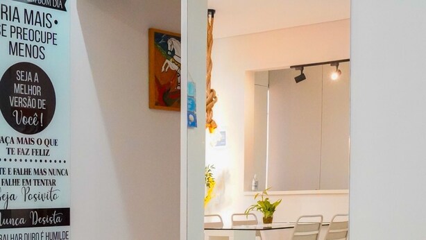 A modern living room with a sofa, cushions, and a coffee table. A large motivational poster is on the wall, and a kitchen area is visible in the background. - Apartment with gourmet balcony, 150m from Enseada beach. (Guarujá)