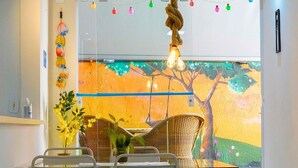 A dining area with wicker furniture, a glass partition, and a colorful mural in the background. - Apartment with gourmet balcony, 150m from Enseada beach. (Guarujá)