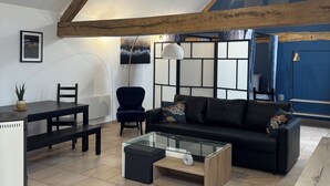 A spacious loft with exposed wooden beams, a black leather sofa, a glass coffee table, and a dining area with a white table and chairs. - Le Merisier (Épieds)
