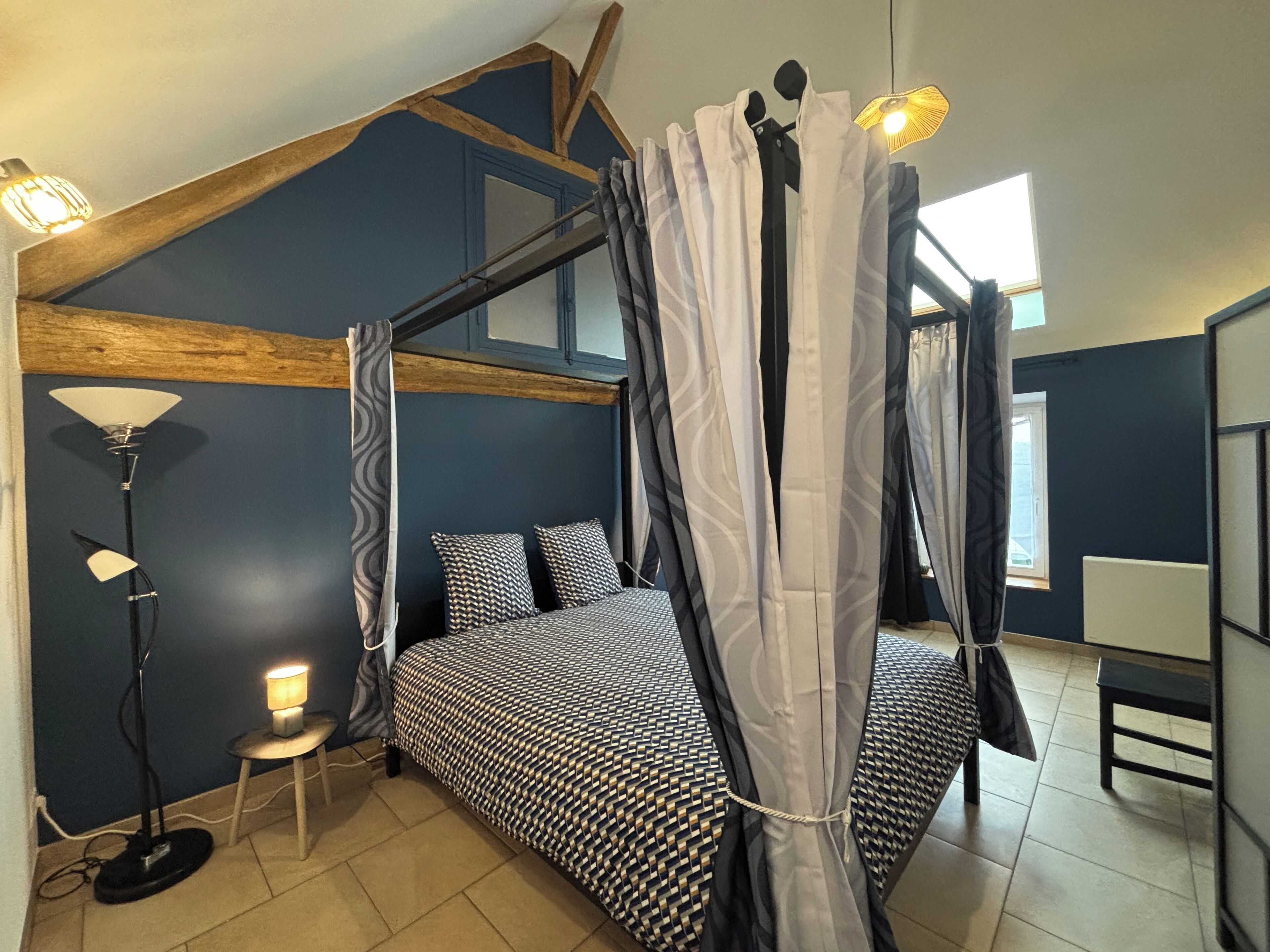 A bedroom with a four-poster bed, a standing lamp, and a view of the sky through the window.