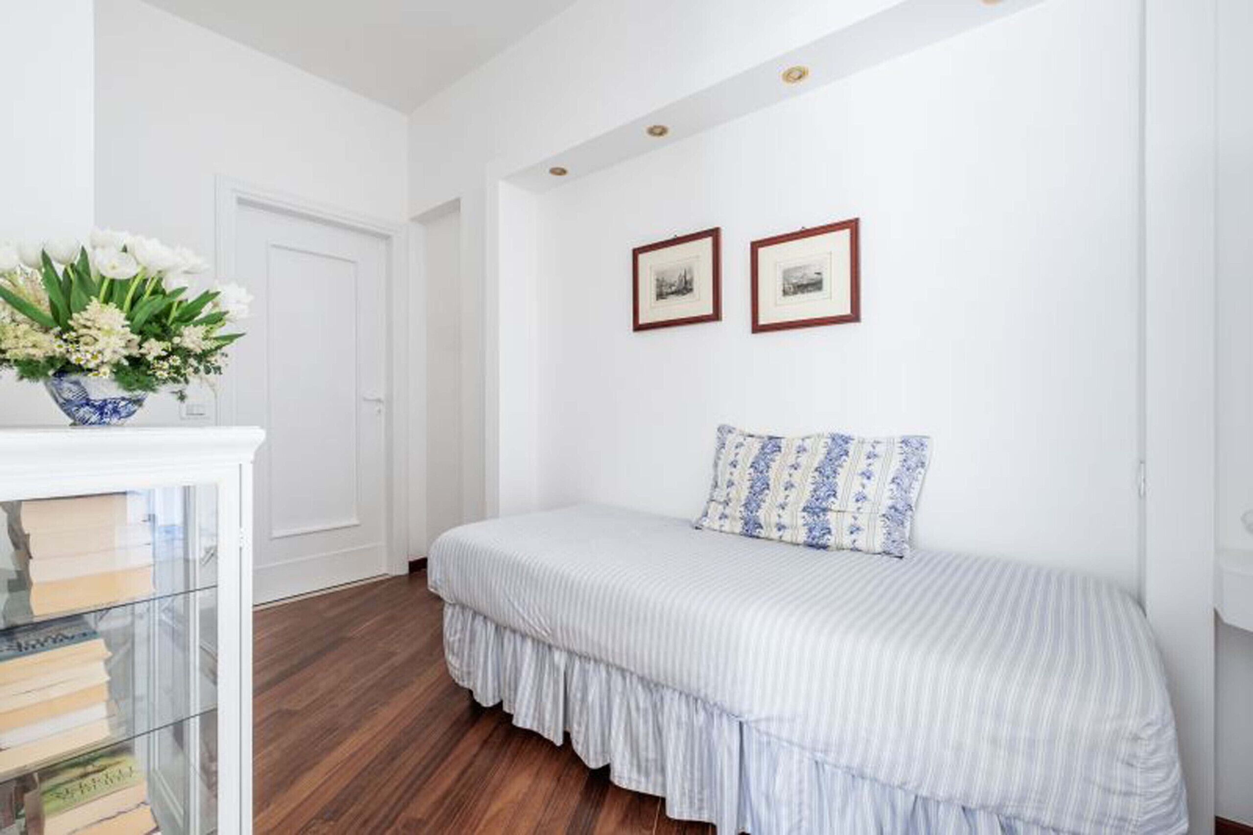 Ein Schlafzimmer mit einem Bett, einem Nachttisch mit Blumenvase und zwei gerahmten Bildern an der Wand.