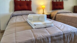 Habitación de hotel con dos camas, cada una con cubrecama acolchado color beige y almohadas rojas. En primer plano, hay una pila de toallas blancas sobre la cama.