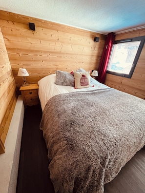 Ein gemütliches Schlafzimmer mit einem Holzkopfteil, einer großen, flauschigen Bettdecke, zwei Kissen, einem Nachttisch mit Lampe und einem Fenster mit roten Vorhängen.