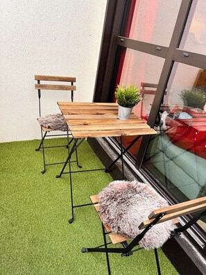 Ein kleiner Sitzbereich im Freien mit Holztisch und -stühlen, einem Blumentopf und einer Glastür zu einem rot gestrichenen Innenraum.