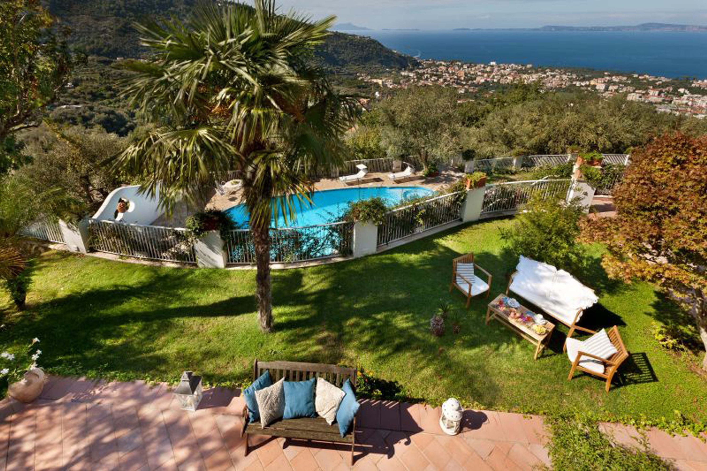A pool surrounded by a fence, a grassy area with a bench, and a view of the sea and hills.