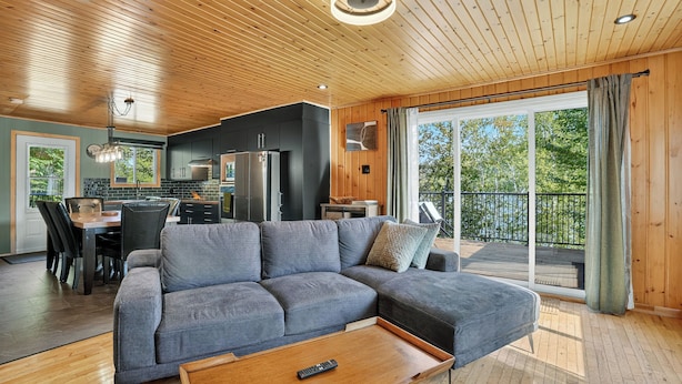 Un salon moderne avec un canapé d’angle gris, un plancher en bois, un coin repas avec une table et des chaises, une cuisine avec des meubles sombres et un réfrigérateur, et un balcon avec vue sur les arbres.
